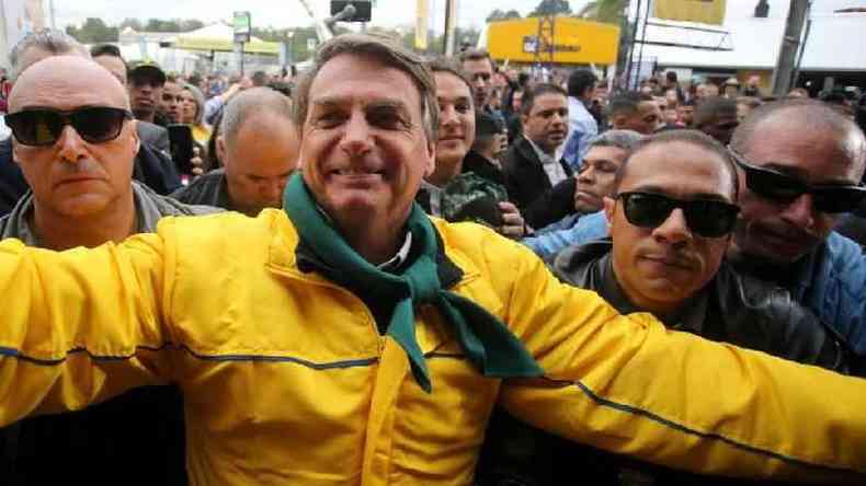 Bolsonaro sorrindo com vestimentas em verde e amarelo rodeado por seguranas e apoiadores nas ruas