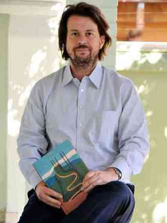 SEntado em varanda, usando cala jeans e camisa azul, Rodrigo Moura segura com as duas mos o livro Lorenzato 