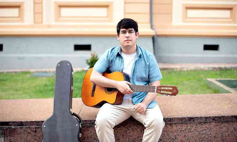 Felipe Bedetti segura o violão, sentado em banco da Praça da Liberdade