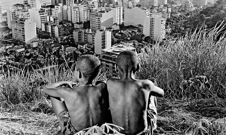 Sentados lado a lado, de costas para a lente, vestindo bermudas, sem camisa e com cabelo quase raspado, dois garotos observam, a partir do morro, a vista de Copacabana