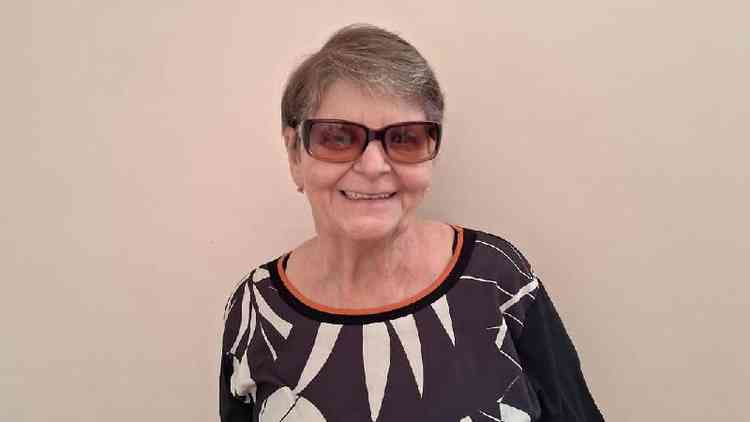 Snia, 67, sorri em frente a uma parede beige; sua pele  branca, ela tem cabelos grisalhos e curtos e usa culos escuros, uma blusa 