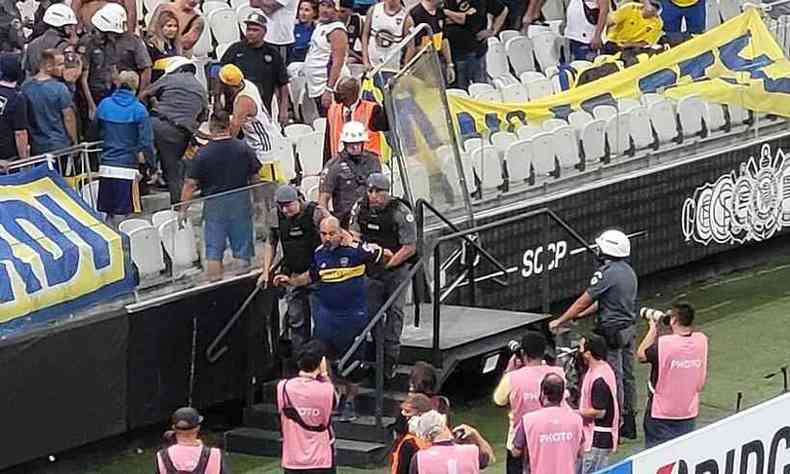 Três policiais guiam um torcedor do Boca Juniors, branco, para fora da arquibancada, que tem uma multidão reunida enquanto observa a movimentação