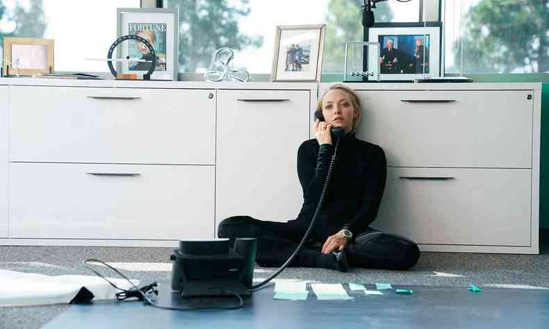 Vestida com cala e blusa pretas, sentada no cho de um escritrio encostada num armrio branco, Amanda Seyfried fala ao telefone em cena de the dropout
