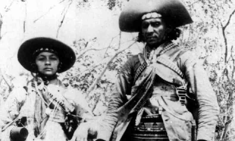 Foto em preto e branco mostra Maria Bonita ao lado de Lampião na caatinga, usando roupas típicas dos cangaceiros 