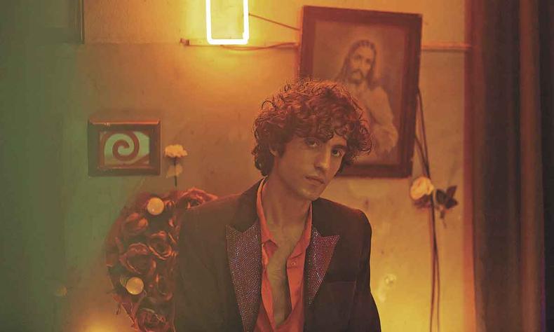 Msico Guto Brant est em ambiente sob penumbra, tendo atrs de si lustre com luz de neon, quadro de Jesus Cristo e flor de plstico presa  parede
