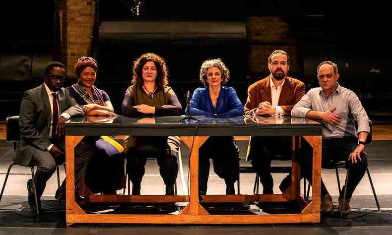 Cinco atores sentados em volta de uma mesa olham para a câmera. Eles participam da peça 'A assembleia São Paulo'