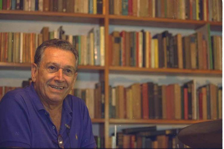 O escritor Fernando Sabino sorri, diante de estante repleta de livros
