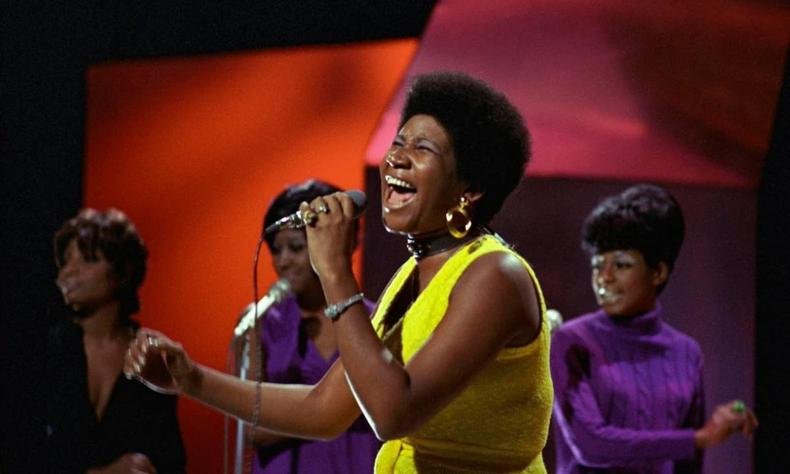 Imagem colorida mostra Aretha Franklin cantando, usando um vestido amarelo 