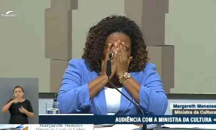 A ministra da Cultura, Margareth Menezes,  uma mulher preta de cabelos cacheados. Ela apoia o rosto nas mos, emocionada