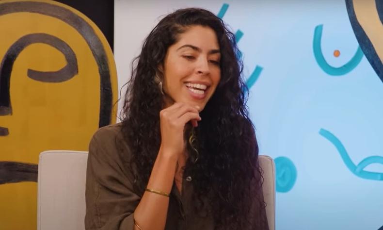 Bela Gil  uma mulher de cabelos longos e cacheados. Ela olha para baixo enquanto leva a mo at o queixo e sorri. Ao fundo, h um mural colorido com amarelo, preto e tons de azul.