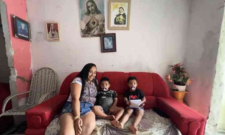 mulher jovem sentada com os filhos gmeos de 3 anos em sof vermelho em frente a parede com retratos religiosos