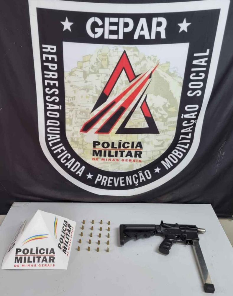 Na foto pode ser visto uma submetralhadora com 20 balas em cima de uma mesa. Logo da Polícia Militar ao fundo