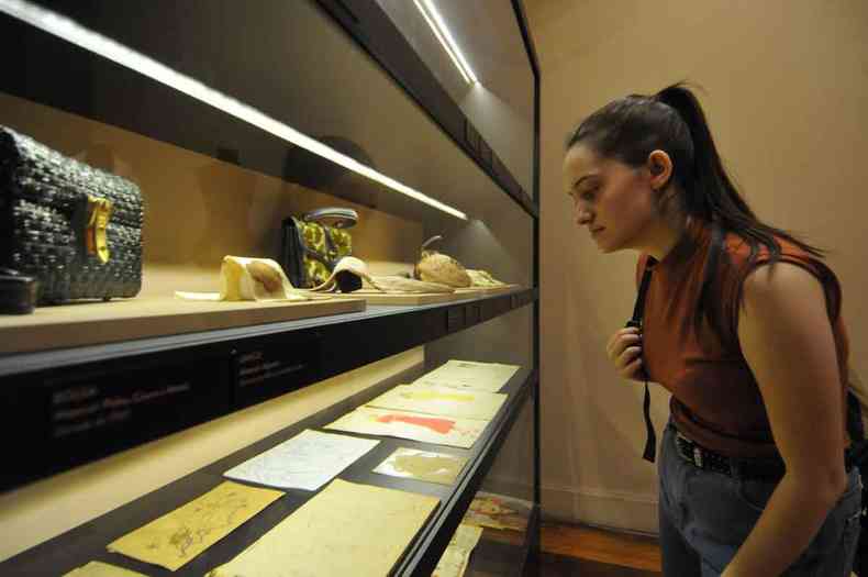Sahra Flrh examina objetos expostos em estante iluminada no Museu da Moda de BH