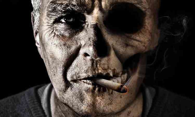 rosto de um homem envelhecido, com cigarro na boca, com metade do rosto normal e a outra uma caveira