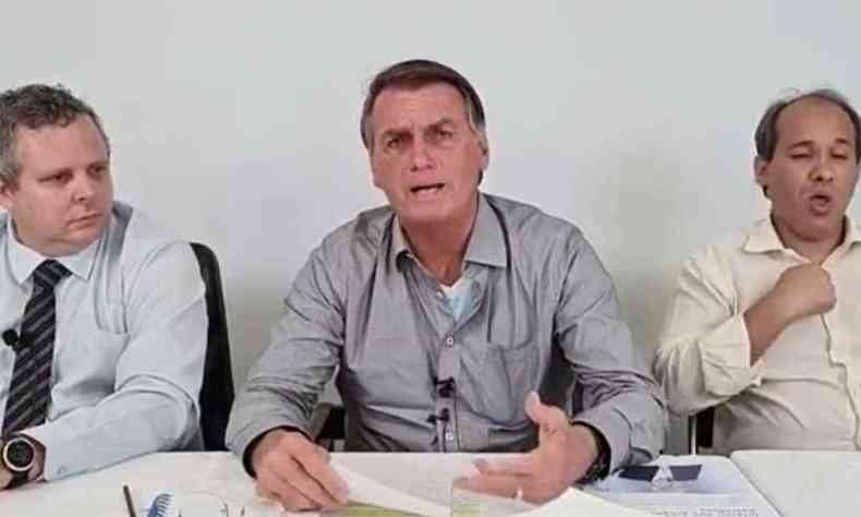 Ao centro, Jair Bolsonaro, um homem branco de cabelos grisalhos e camisa social cinza gesticula com um papel em mos. Ao seu lado, esto dois assessores, ambos brancos e de camisa social. Um deles, gesticula em lngua de sinais.