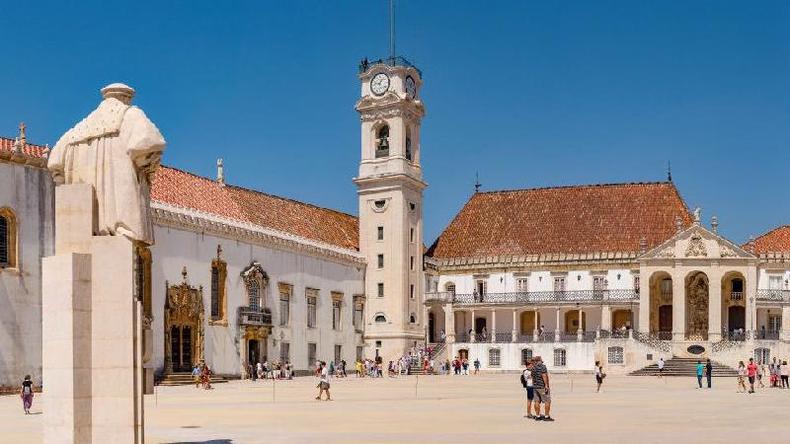 Torre da Universidade de Coimbra diante de ptio com pessoas caminhando em um dia de sol; h uma esttua de costas