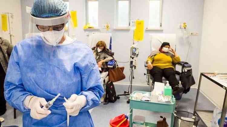 Profissional de sade com roupa de proteo manipulando medicamentos quimioterpicos, com pacientes de cncer em tratamento ao fundo