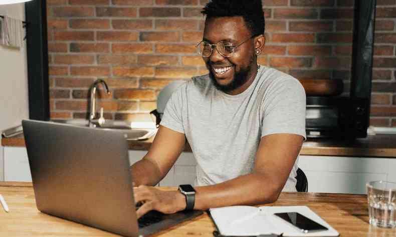 Homem negro sorrindo, sentado  mesa, usando laptop