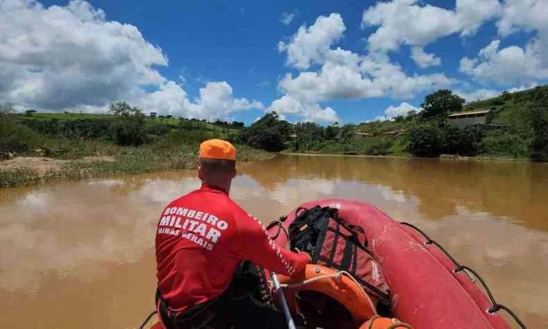 Um homem do corpo de bombeiros, blusa vemelha e capacete laranja em cima de um barco atravessando um rio de cor marrom, buscando vtimas de afogamentos