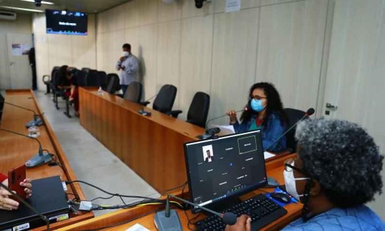 Iza Lourena e Maca Evaristo, ambas mulheres negras, sentam-se em uma mesa comprida usando mascaras descartveis. Em frente  Maca, h uma tela de computador aberta em uma reunio online