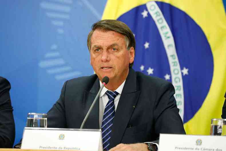 Presidente Jair Bolsonaro fala ao microfone; ao fundo uma bandeira do Brasil