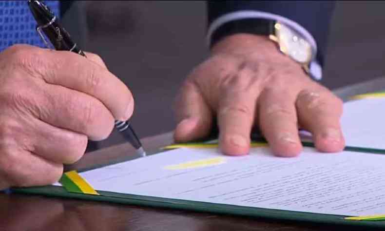 Lula est assinando a sano da Lei que tipifica injria racial como crime de racismo. A imagem foca em suas mos: uma se apoia na mesa e a outra segura a caneta.