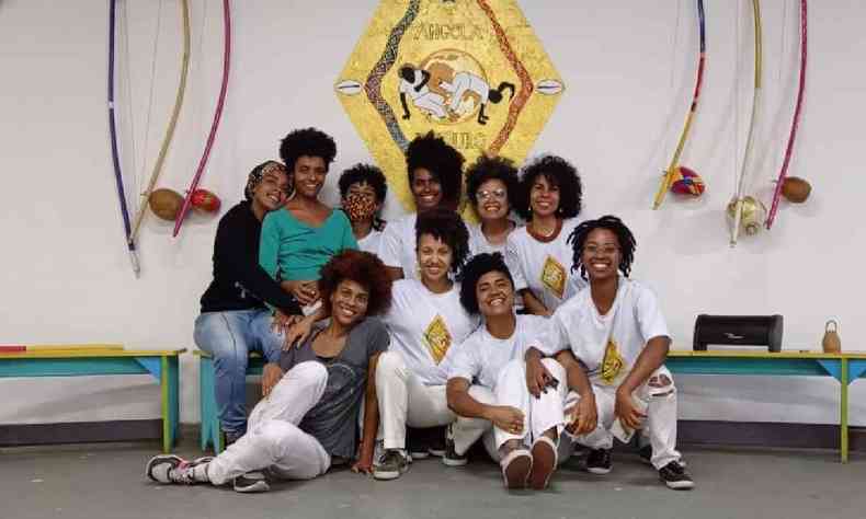 Mulheres em roupas de capoeira sentadas em frente ao logo do Coletivo Cultural Angola de Ouro