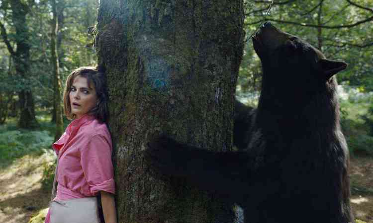 Atriz Keri Russell com a cara assustada est encostada a uma rvore. Do outro lado do tronco, imenso urso negro olha para cima, em cena do filme O urso do p branco 
