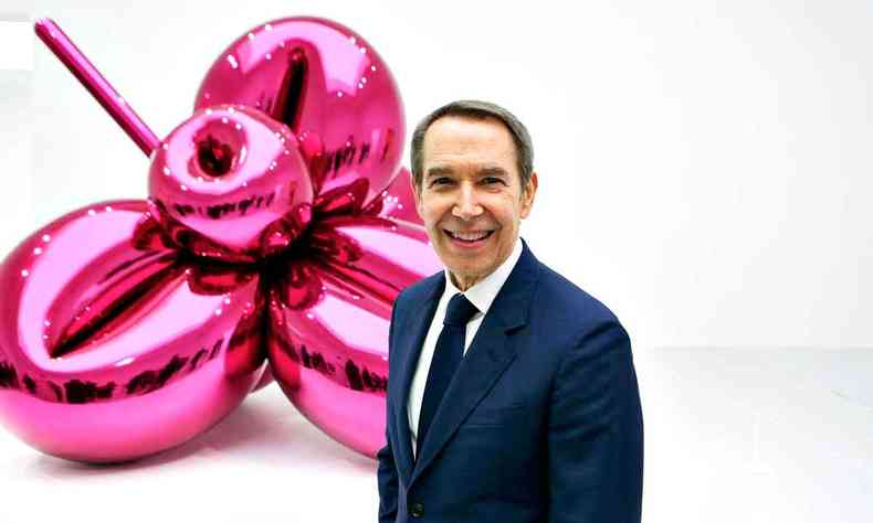 O artista plstico Jeff Koons sorri e ao fundo v-se escultura assinada por ele, no tom rosa escuro, semelhante a um balo em forma de flor