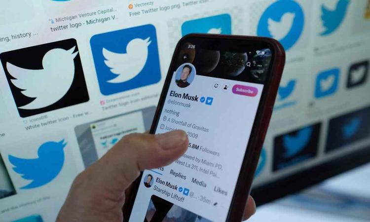 Pessoa segura o celular diante de um painel com vrias logomarcas do Twitter; na tela, o perfil de Elon Musk