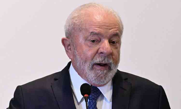 Lula com camisa branca, terno e gravata pretos, falando em um microfone em um fundo branco