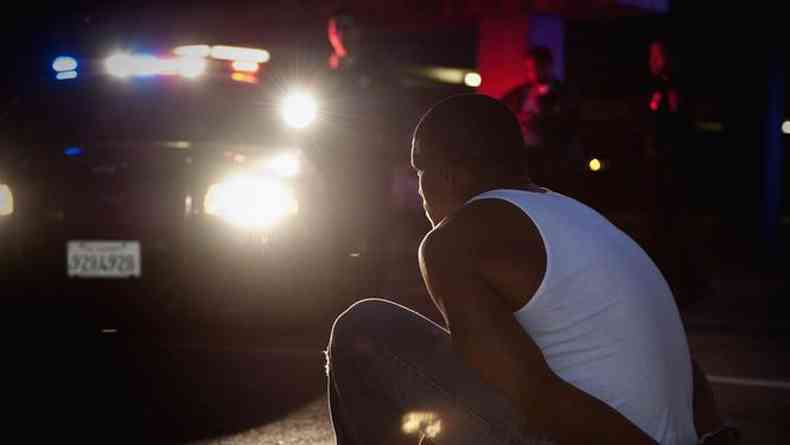 Homem negro sentado e algemado no meio-fio, observado por policiais ao lado de viatura a noite