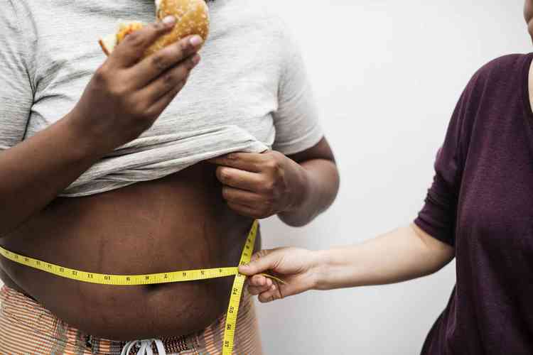 Mulher medindo barriga de homem obeso que est segurando um sanduche