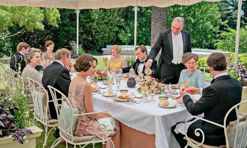 Em refeio no jardim, pessoas vestidas luxuosamente esto  mesa, servidas por empregados trajados com roupas formais, durante cena do filme Downton Abbey II - Uma nova era