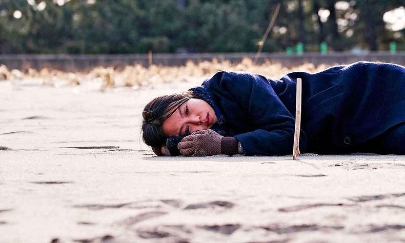 Atriz deitada na areia da praia com semblante triste em cena de filme