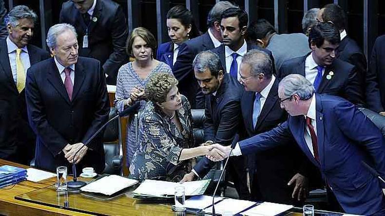 Dilma e Eduardo Cunha apertando as mos diante da mesa da Cmara dos Deputados, rodeados por outras pessoas