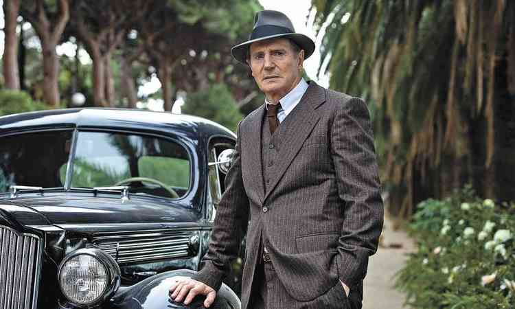 O ator Liam Neeson, de terno e chapu, apoiado em carro antigo, em cena de Sombras de um crime
