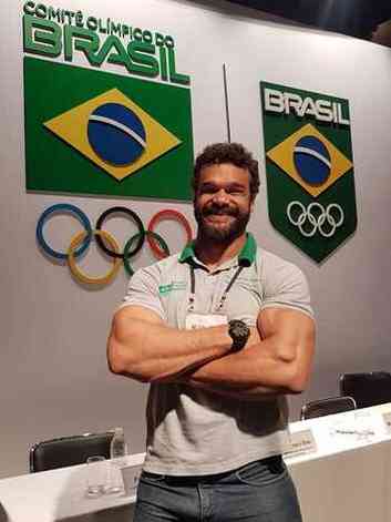 Homem branco, barbudo, atleta, com blusa branca e cala jeans de braos cruzados sorrindo para a foto na frente de uma mesa do comit olimpico brasileiro