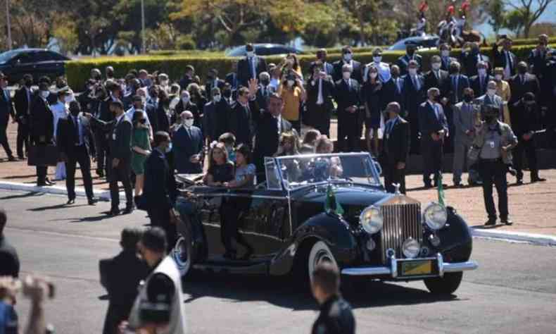 No centro da imagem Rolls Royce com presidente jair bolsonaro e crianas, ao redor do veculo pessoas observam a passagem do presidente. Ele esta em pe no carro acenando para as pessoas