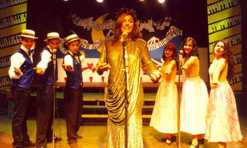 A atriz Kenya Costa, de vestido dourado, canta no microfone, ao centro, tendo ao fundo coro formado por três rapazes e três moças, na peça Nas ondas do rádio