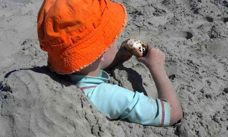 criana enterrada na areia na praia, de chapu e comendo um bolinho